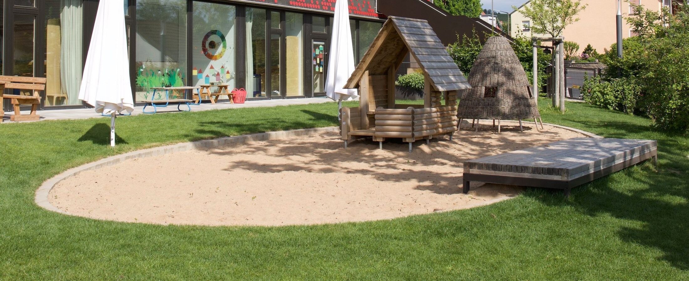Garten der Kita mit Sankasten, Spielhaus und dem Gebäude im HIntergrund